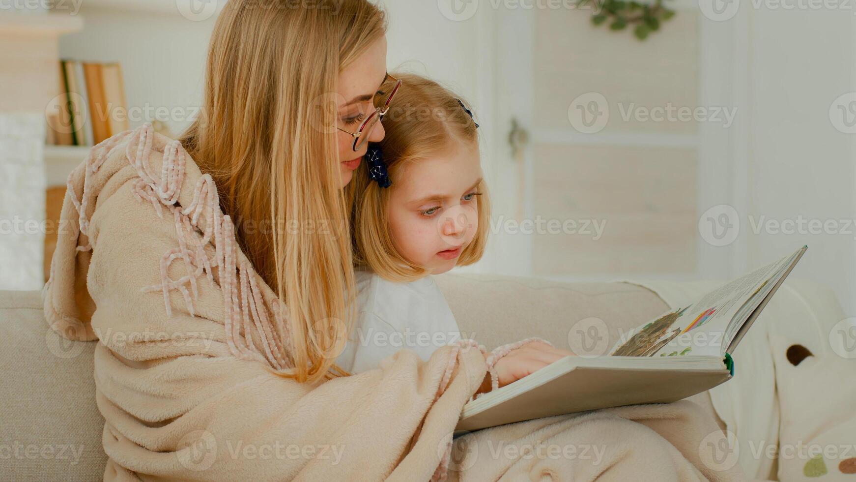 poco piccolo caucasico figlia bambino ragazza ragazzo bambino con madre nel vivente camera lettura letteratura guardare a foto album mamma leggere libro mostrando immagini spiegare a divano divano famiglia elementare apprendimento