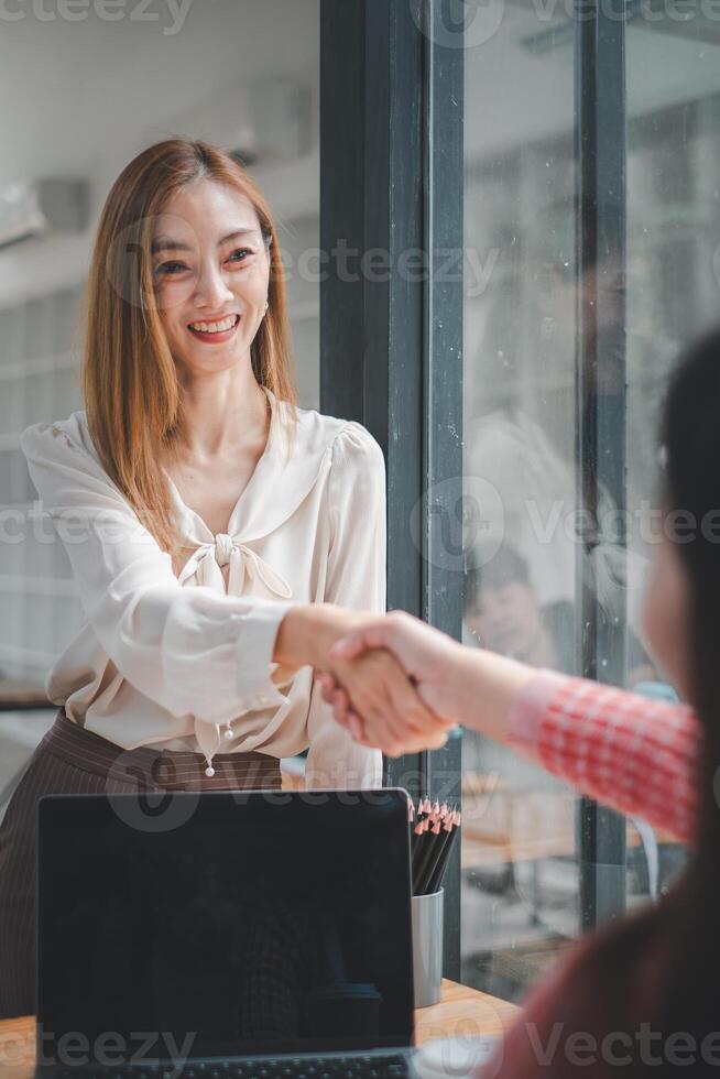 Due sorridente donne d'affari tremante mani nel accordo durante un' riuscito ufficio incontro, con un' il computer portatile su il tavolo. foto