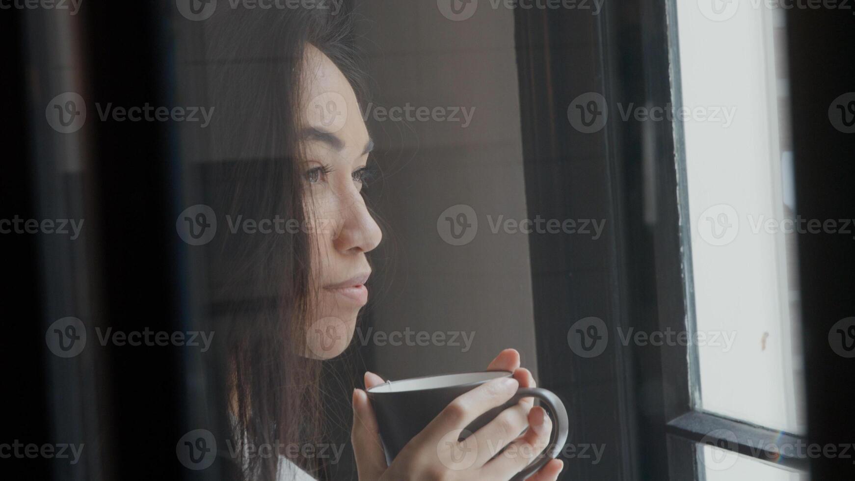 giovane donna di razza mista cammina tenendo la tazza con entrambe le mani, si avvicina alla finestra, beve un sorso, guarda contemplativamente attraverso la finestra foto