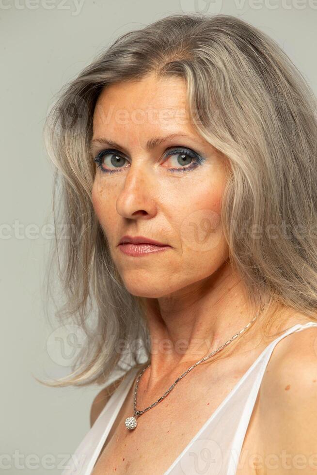 bellissimo dai capelli grigi cinquanta anno vecchio donna guardare a telecamera isolato su grigio sfondo. maturo vecchio signora vicino su ritratto. il viso di un invecchiamento donna con rughe e imperfezioni. foto