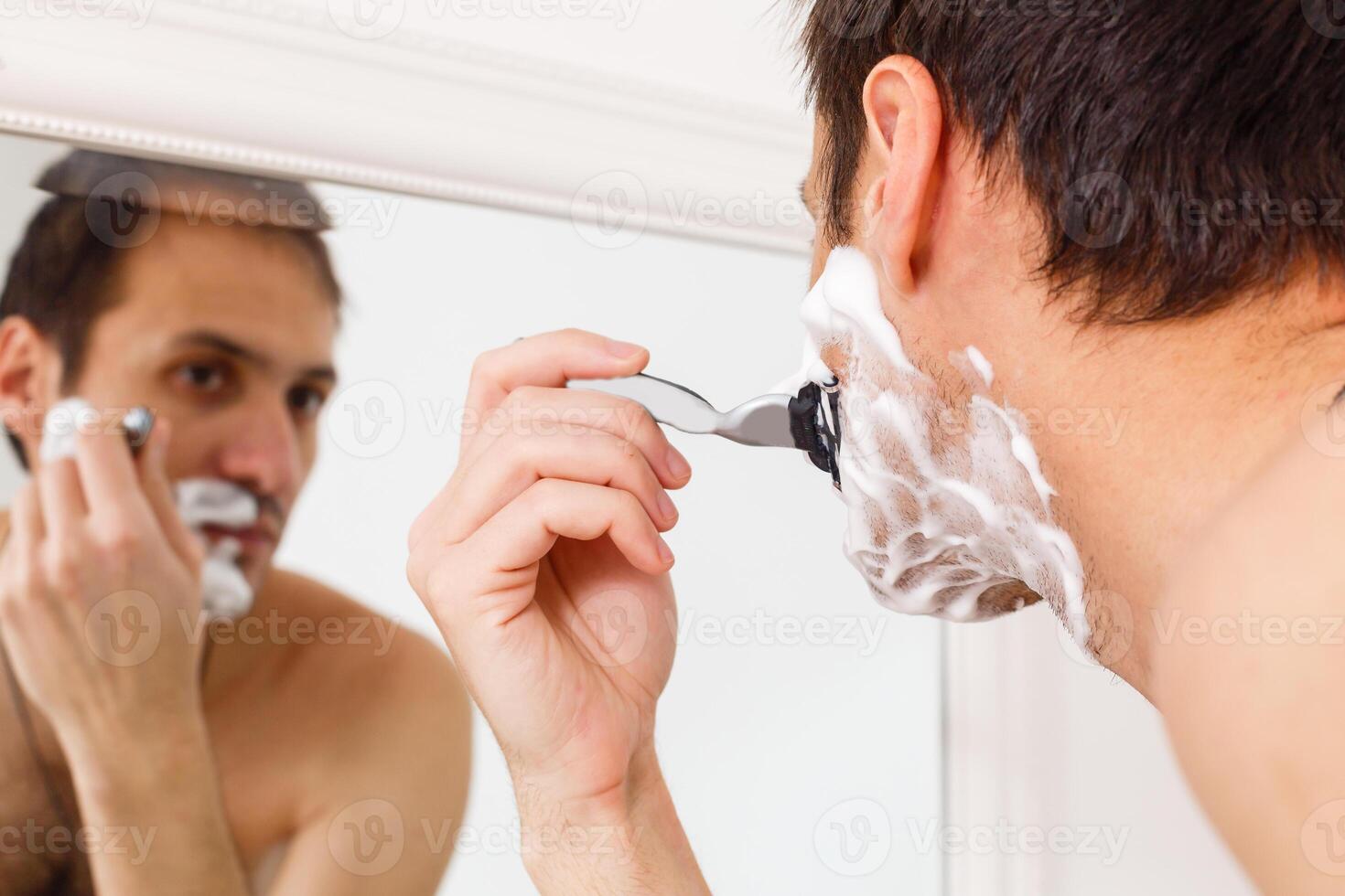 giovane uomo rasatura nel il bagno. lui è passaggio il rasoio per il barba mentre esso sembra a il specchio foto