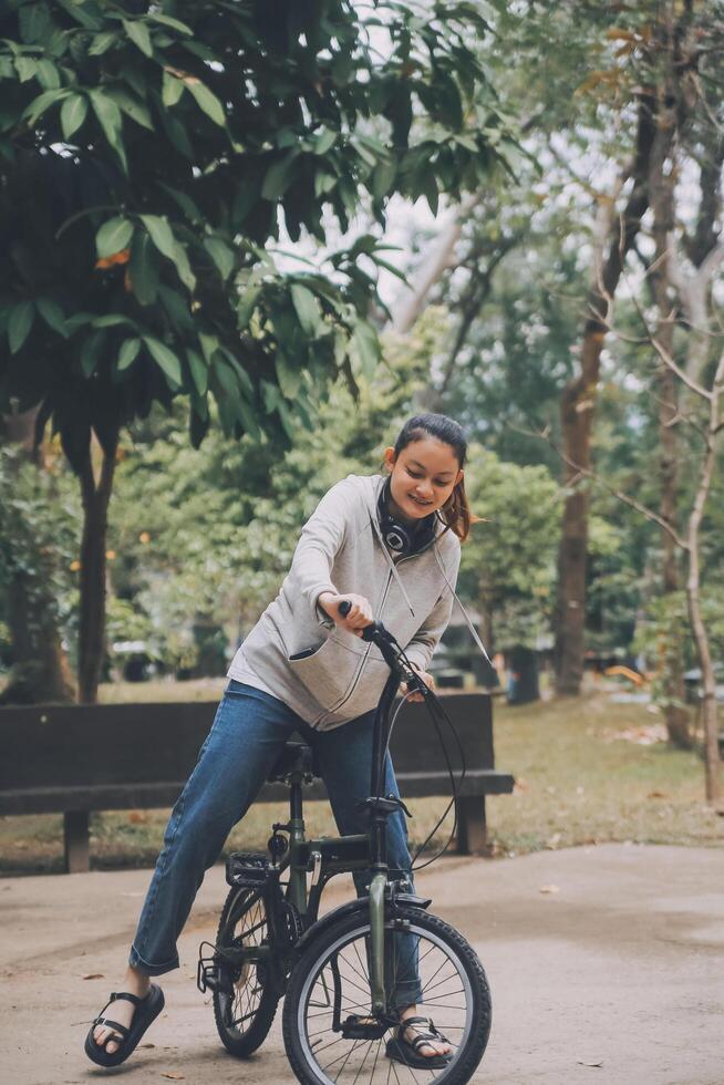 contento asiatico giovane donna camminare e cavalcata bicicletta nel parco, strada città sua sorridente utilizzando bicicletta di trasporto, eco amichevole, persone stile di vita concetto. foto