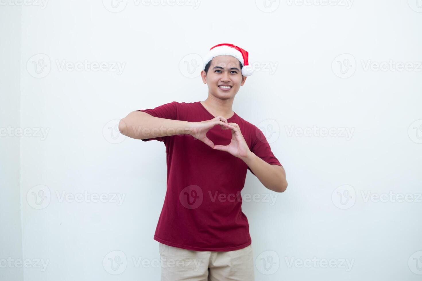 giovane asiatico uomo indossare un' Santa Claus cappello esprimendo amore di formatura un' cuore simbolo con il suo mani isolato di un' bianca sfondo per visivo comunicazione foto