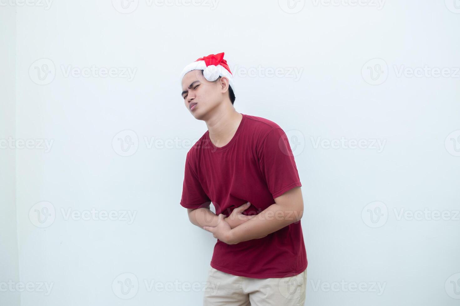 giovane asiatico uomo indossare un' Santa Claus cappello esprimendo mal di stomaco di Tenere il suo stomaco nel dolore isolato di un' bianca sfondo per visivo comunicazione foto