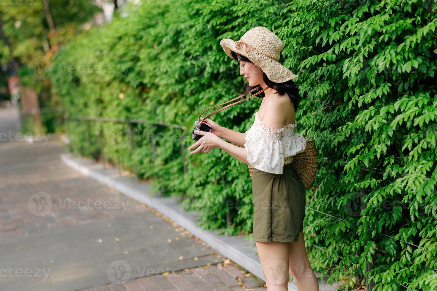 ritratto di asiatico giovane donna viaggiatore con tessitura cappello e cestino e un' telecamera su verde pubblico parco natura sfondo. viaggio viaggio stile di vita, mondo viaggio esploratore o Asia estate turismo concetto. foto