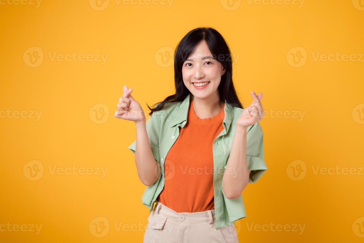 bello giovane asiatico donna nel sua 30s mostrando un' mini cuore dito gesto, esprimendo affetto con dolce Sorridi. concetto di corpo linguaggio come lei pose in casa indossare un arancia camicia e verde Maglione. foto