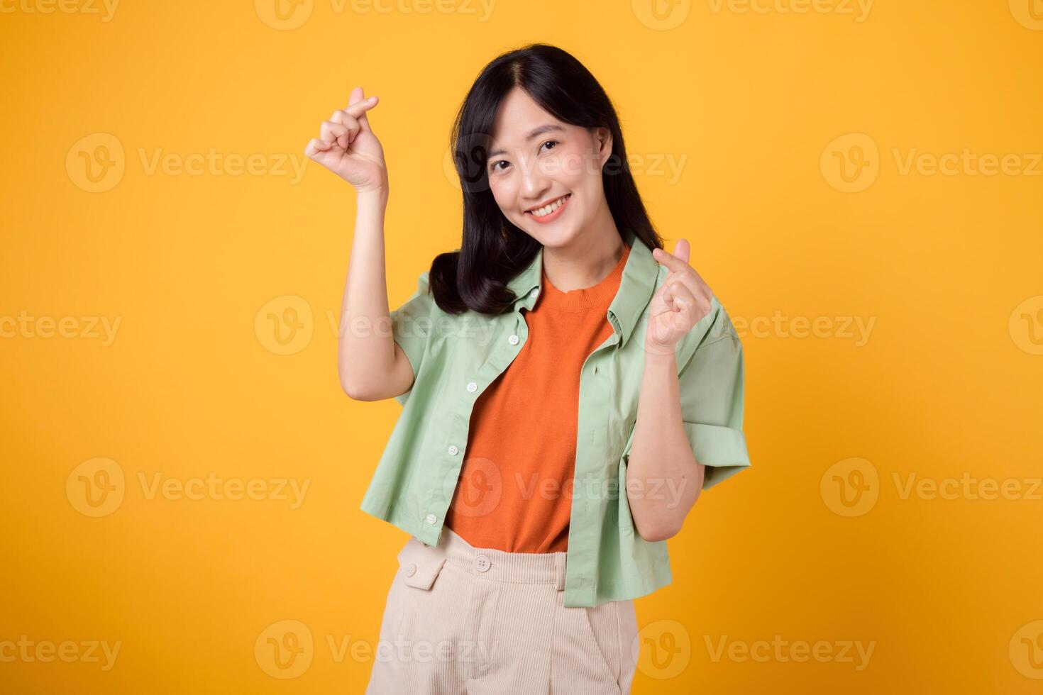 giovane asiatico donna nel sua 30s vestito nel un arancia camicia e verde Maglione. sua mini cuore gesto e dolce Sorridi parlare volumi di affetto e felicità. corpo linguaggio concetto. foto