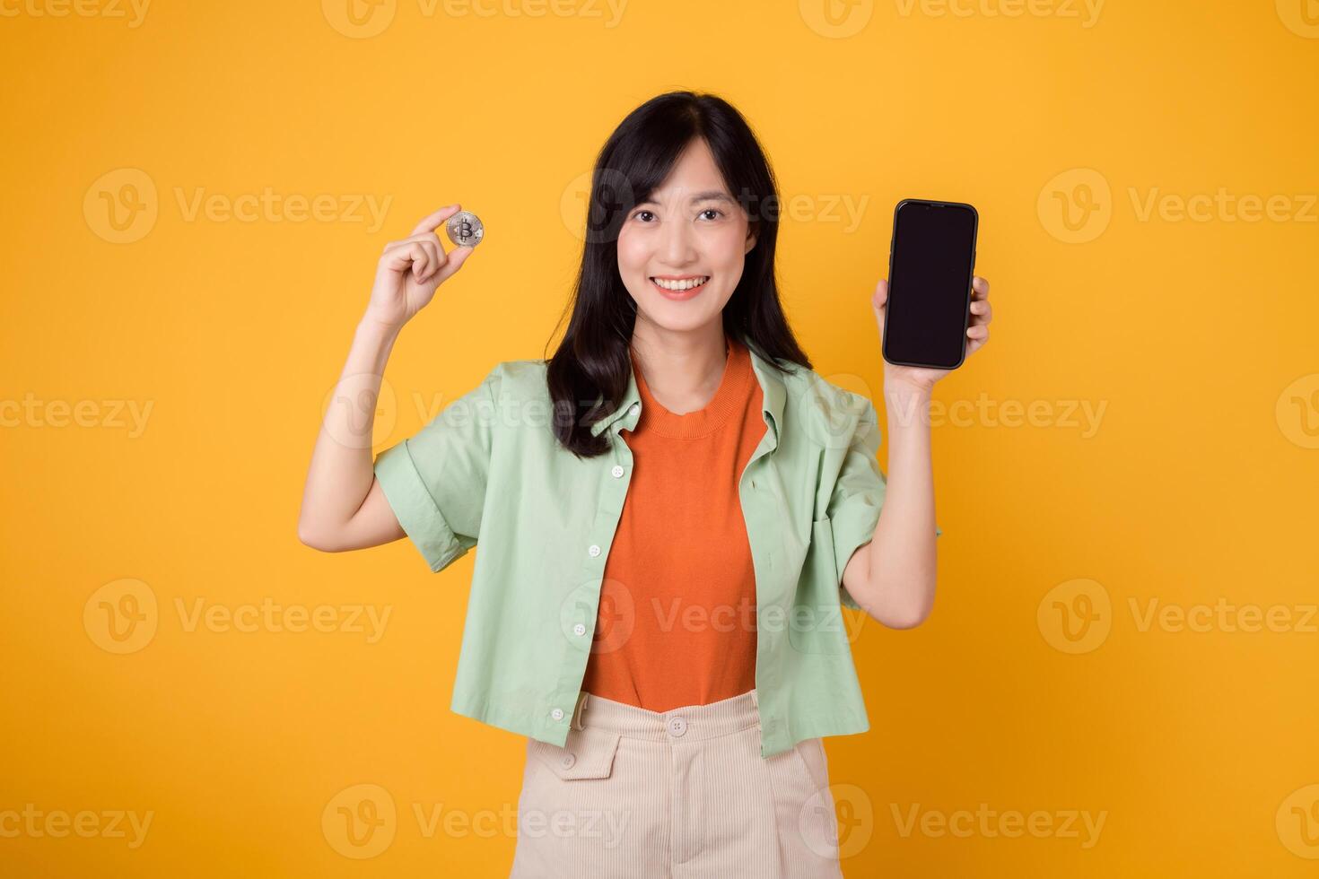 il futuro di finanza con un' travolgente giovane asiatico donna nel sua anni '30, elegantemente vestito nel arancia camicia e verde Maglione, utilizzando smartphone schermo e crypto moneta moneta su giallo sfondo. foto