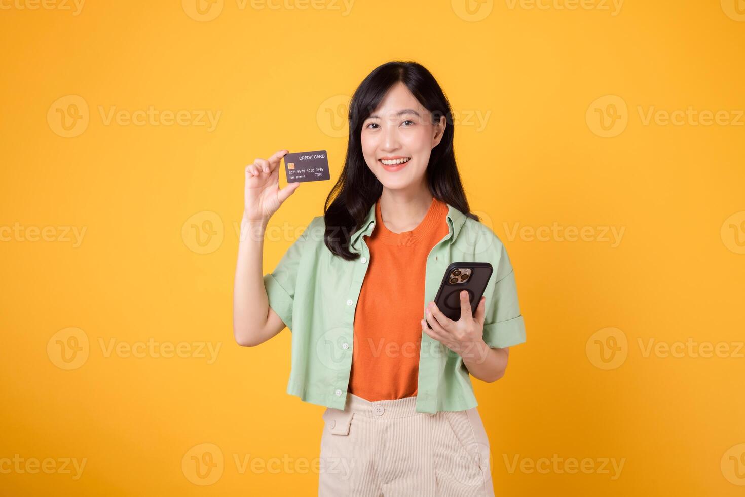 scoprire il gioia di in linea shopping con allegro giovane asiatico donna nel sua anni '30, indossare arancia camicia e verde Maglione, utilizzando smartphone per mostrare credito carta su giallo studio sfondo. mobile concetto. foto