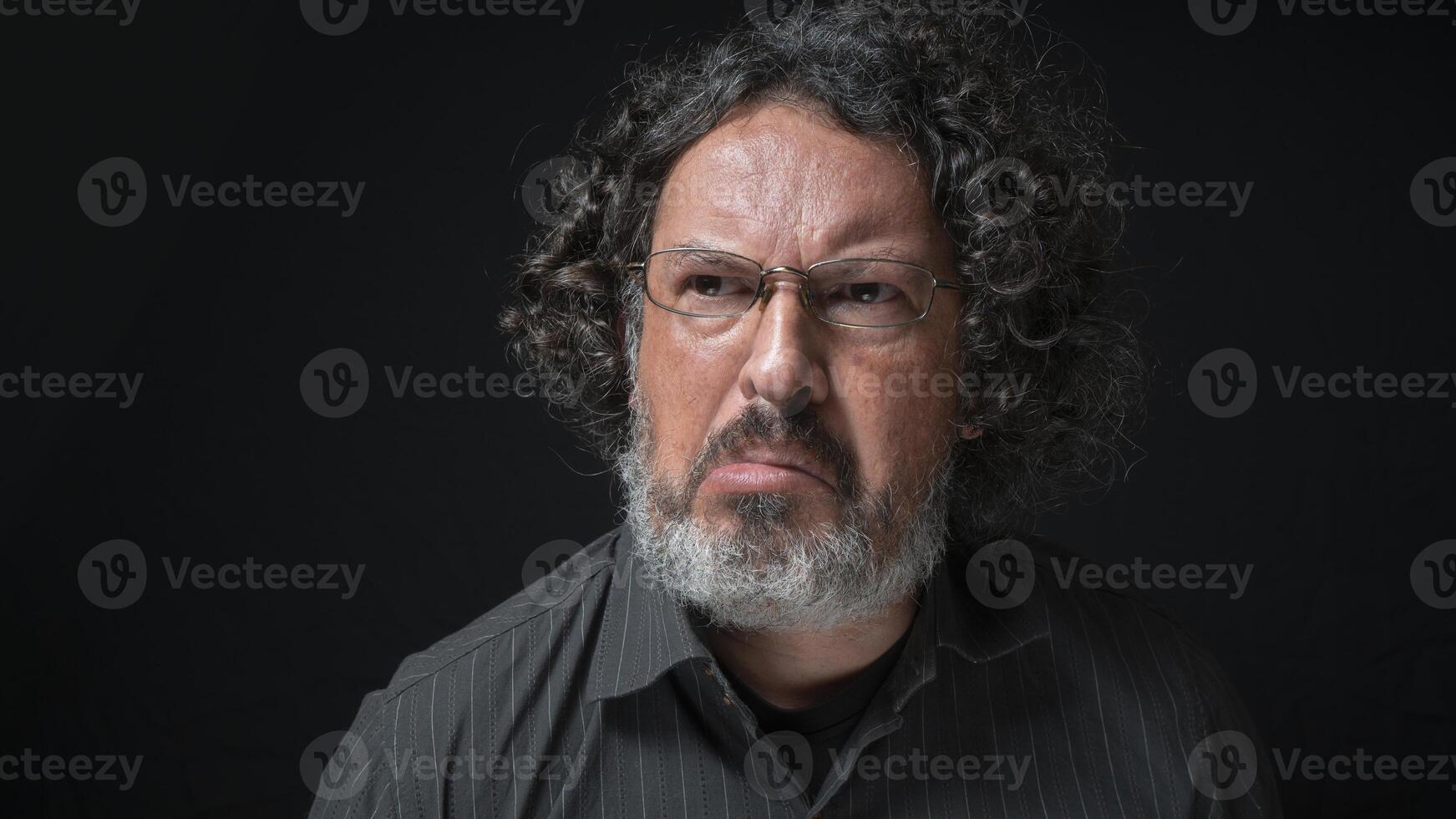 uomo con bianca barba e nero Riccio capelli con frustrato espressione, smorfie con bocca, indossare nero camicia contro nero sfondo foto