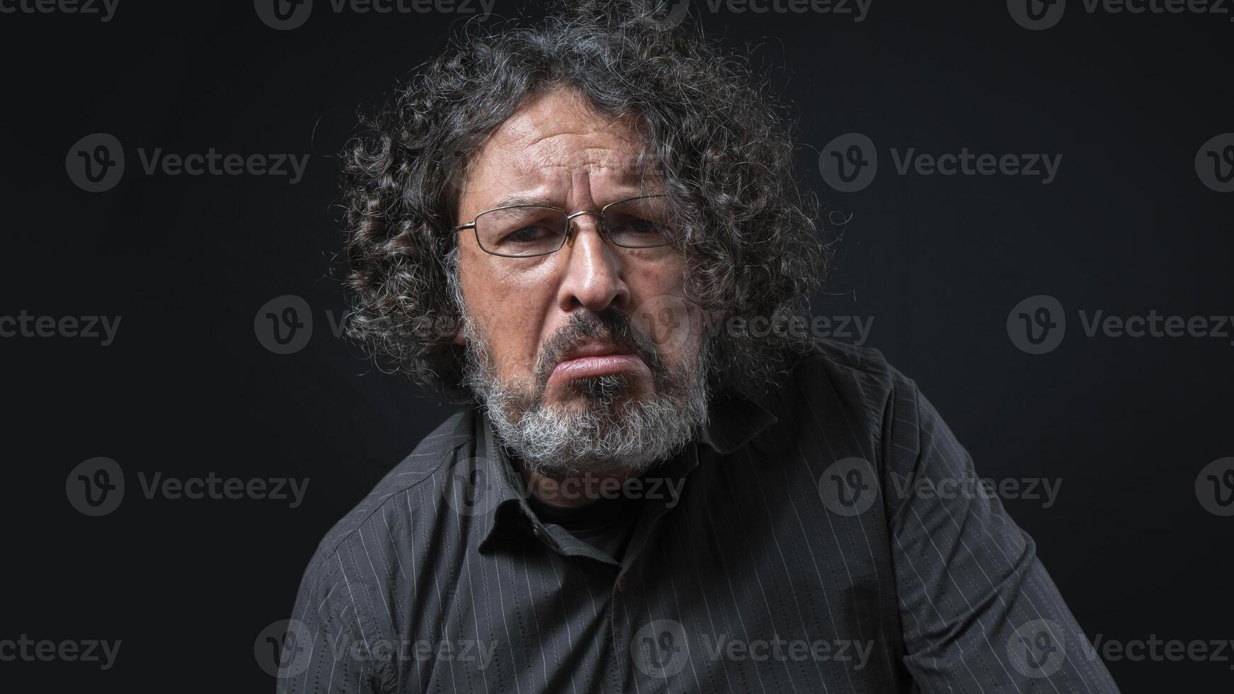 uomo con bianca barba e nero Riccio capelli con triste espressione, indossare nero camicia contro nero sfondo foto