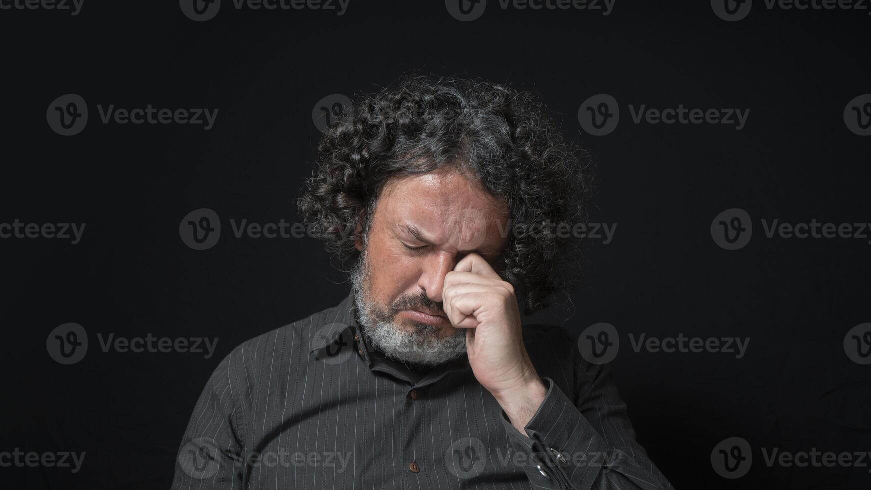 uomo con bianca barba e nero Riccio capelli con triste espressione, occhi chiuso e testa fuori uso, indossare nero camicia contro nero sfondo foto