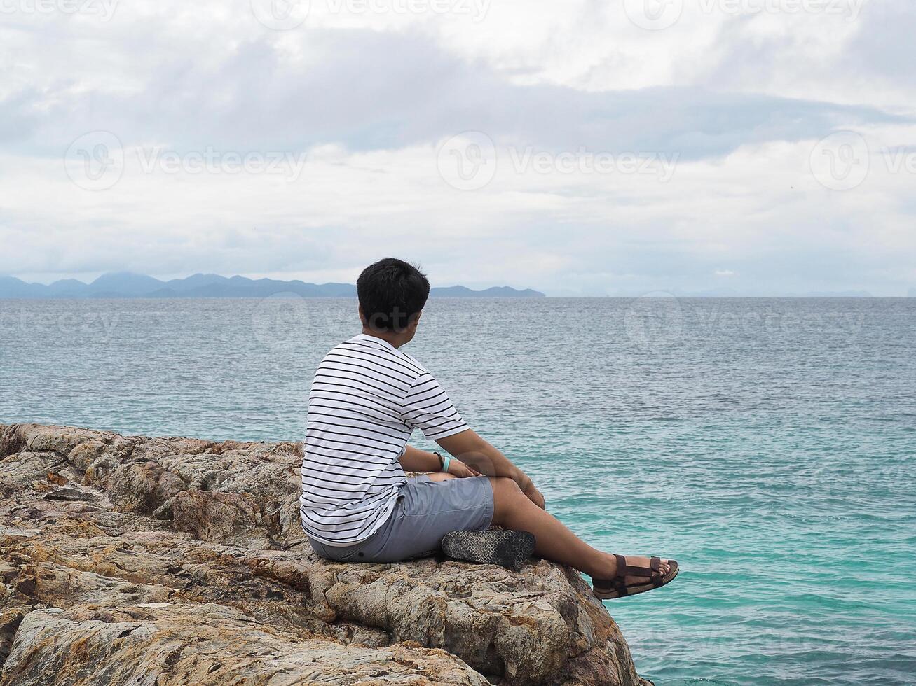 indietro Visualizza di donna corto capelli seduta su un' roccioso riva e guardare a il mare foto