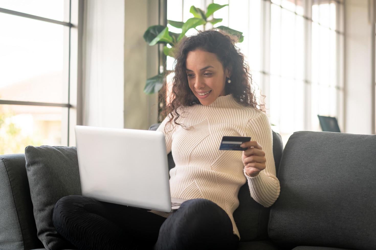 donna latina che utilizza laptop e mano che tiene la carta di credito per lo shopping sul divano foto