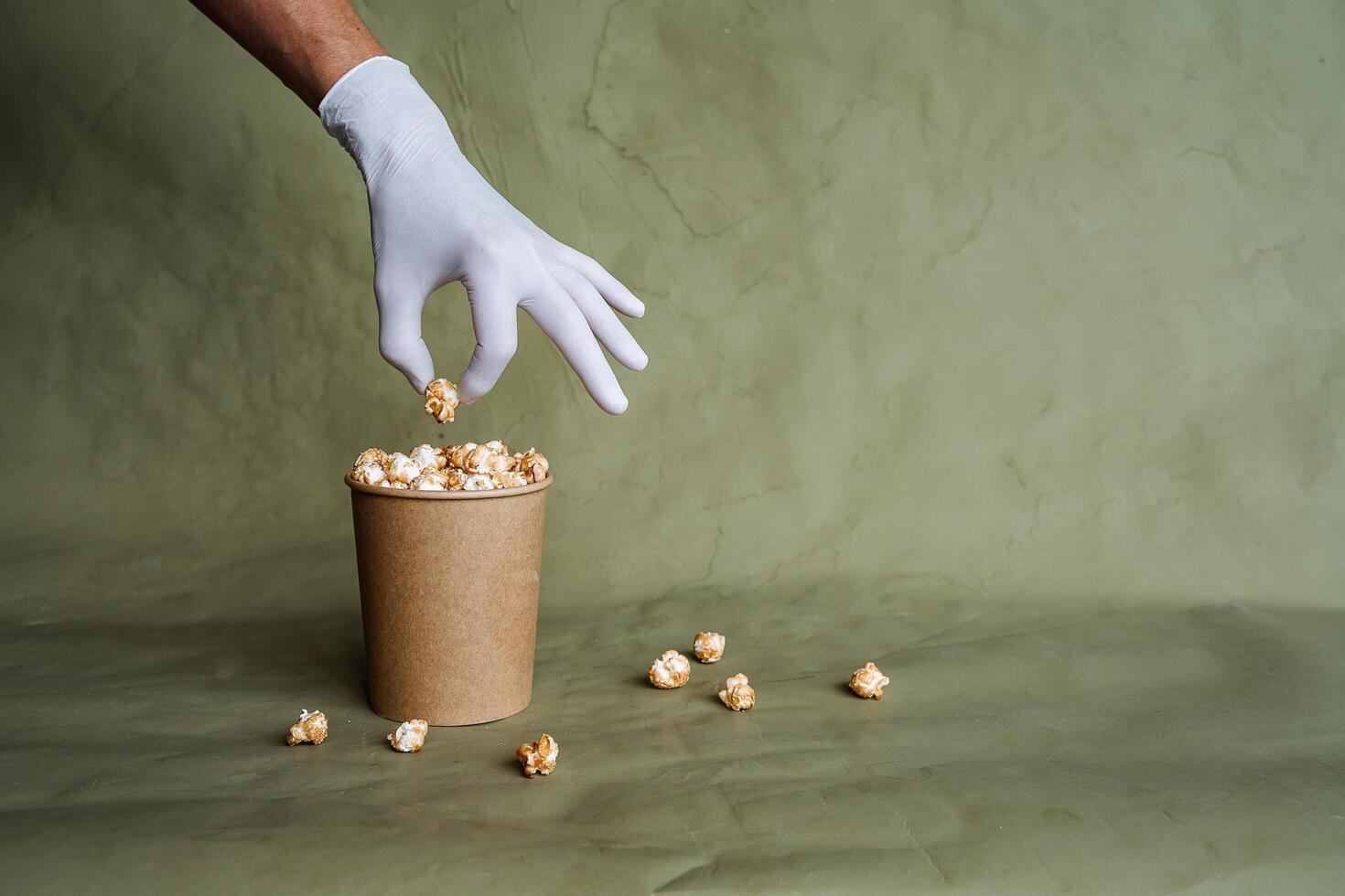 mano nel un' bianca guanto raggiunge per Popcorn, prendere cibo nel protettivo guanti, hold un' pezzo di cibo con Due dita, un' pieno scatola di Popcorn, sicurezza a il veloce cibo distribuzione, minimo concetto foto