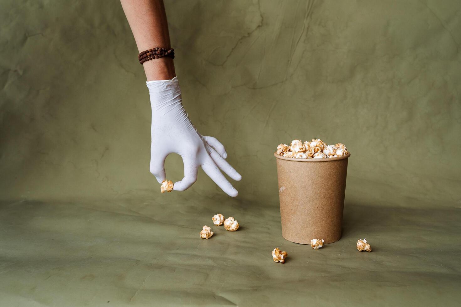 mano nel un' bianca guanto raggiunge per Popcorn, prendere cibo nel protettivo guanti, hold un' pezzo di cibo con Due dita, un' pieno scatola di Popcorn, sicurezza a il veloce cibo distribuzione, minimo concetto foto