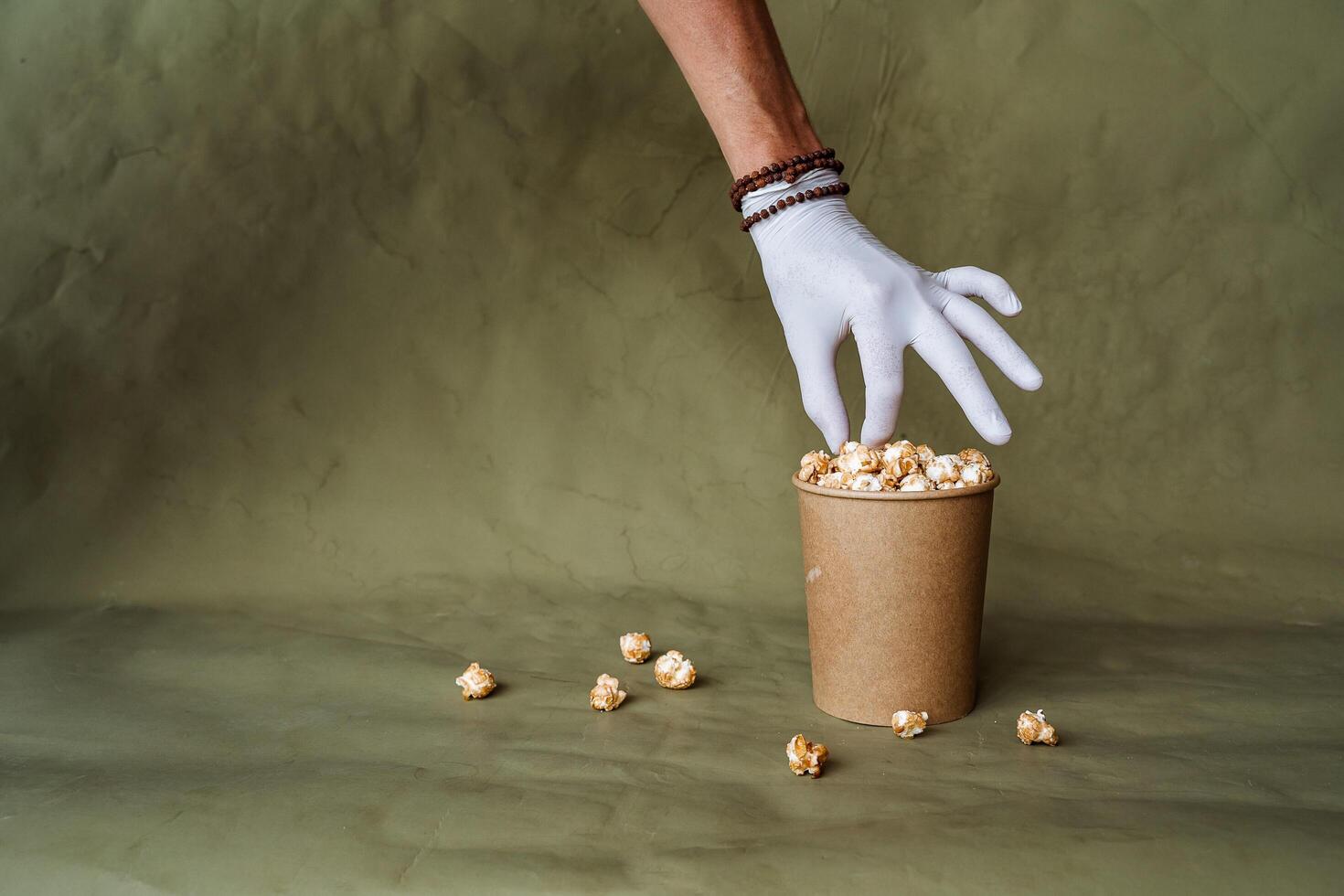 mano nel un' bianca guanto raggiunge per Popcorn, prendere cibo nel protettivo guanti, hold un' pezzo di cibo con Due dita, un' pieno scatola di Popcorn, sicurezza a il veloce cibo distribuzione, minimo concetto foto