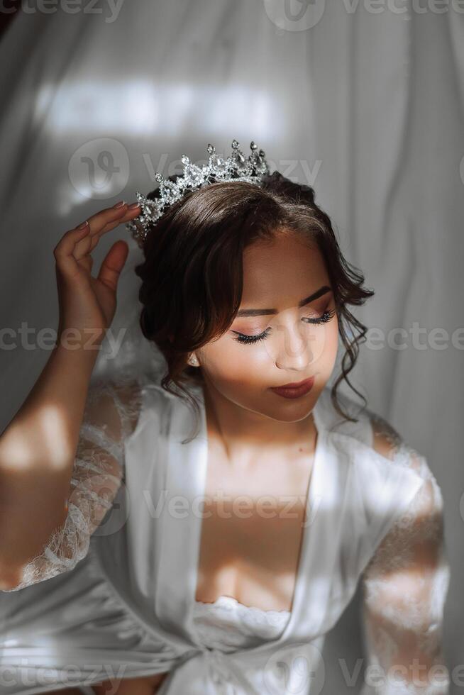 un' bellissimo brunetta sposa con un' tiara nel sua capelli è ottenere pronto per il nozze nel un' bellissimo accappatoio nel boudoir stile. avvicinamento nozze ritratto, foto. foto