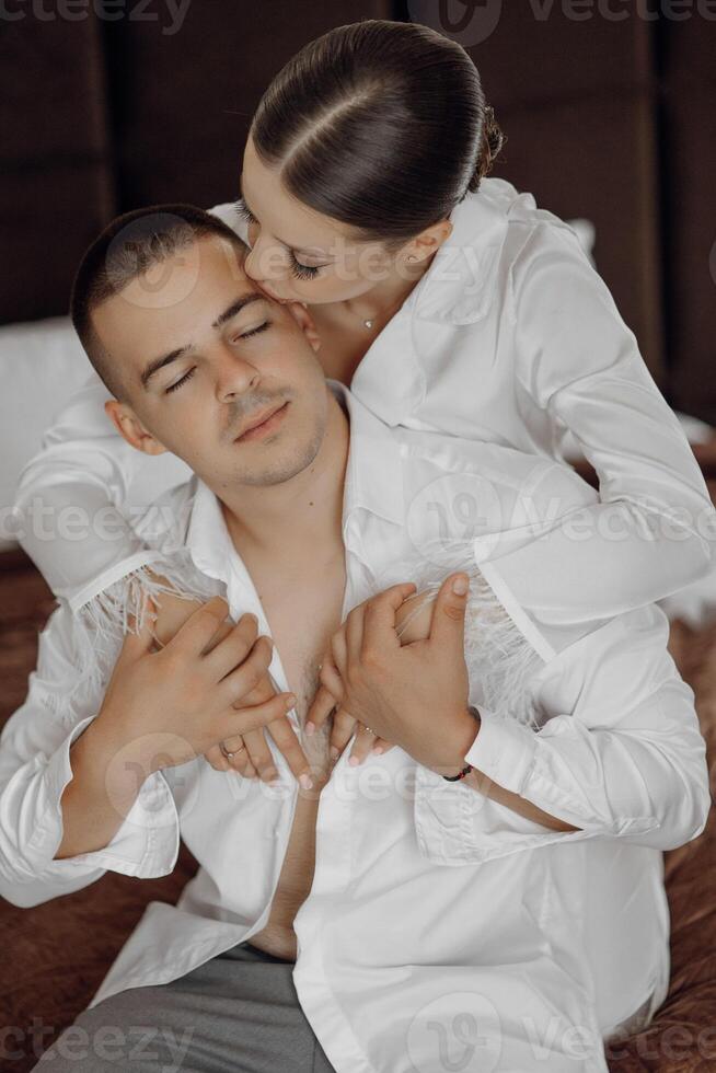 gioioso giovane sposa nel bianca pigiama abbracciare il le spalle di bello sposo nel bianca sbottonato camicia seduta insieme nel moderno Hotel camera prima nozze cerimonia foto