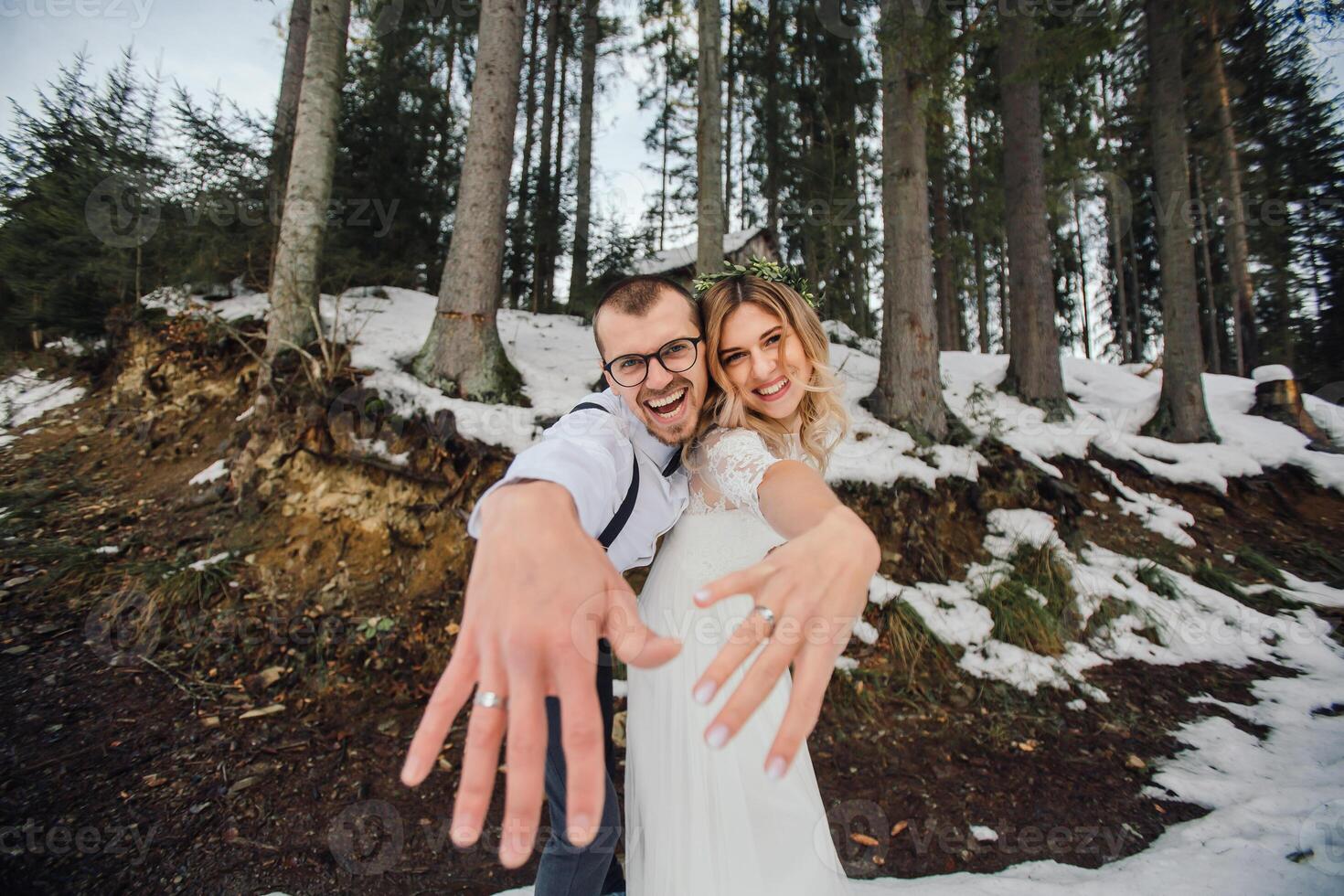 un' giovane coppia, il sposa e sposo, sorridendo mostrare via loro nozze anelli contro il sfondo di alto alberi. avere un' bene tempo ridendo. inverno nozze foto