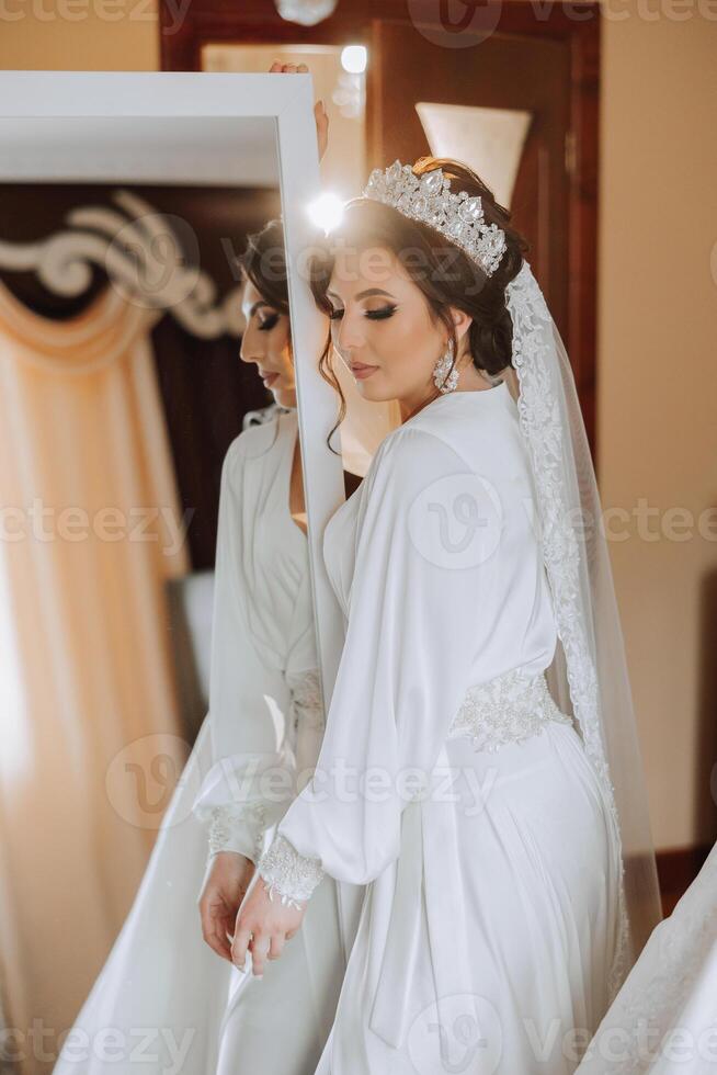 ritratto di un' lussuoso sposa nel boudoir stile vicino un' specchio con riflessione. preparazione il sposa per il nozze nel il mattina nel un' bianca veste. foto