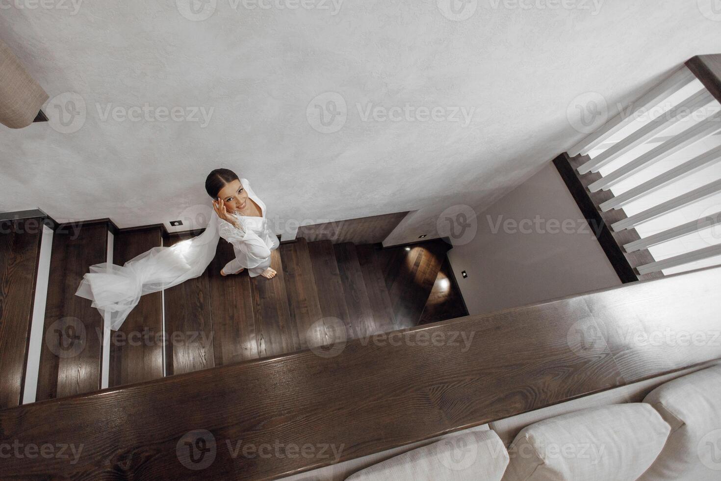 un' bellissimo e fiducioso sposa nel pigiama con un' lungo velo passeggiate su il di legno Marrone le scale nel il Hotel camera. nozze concetto. mattina di il sposa. foto