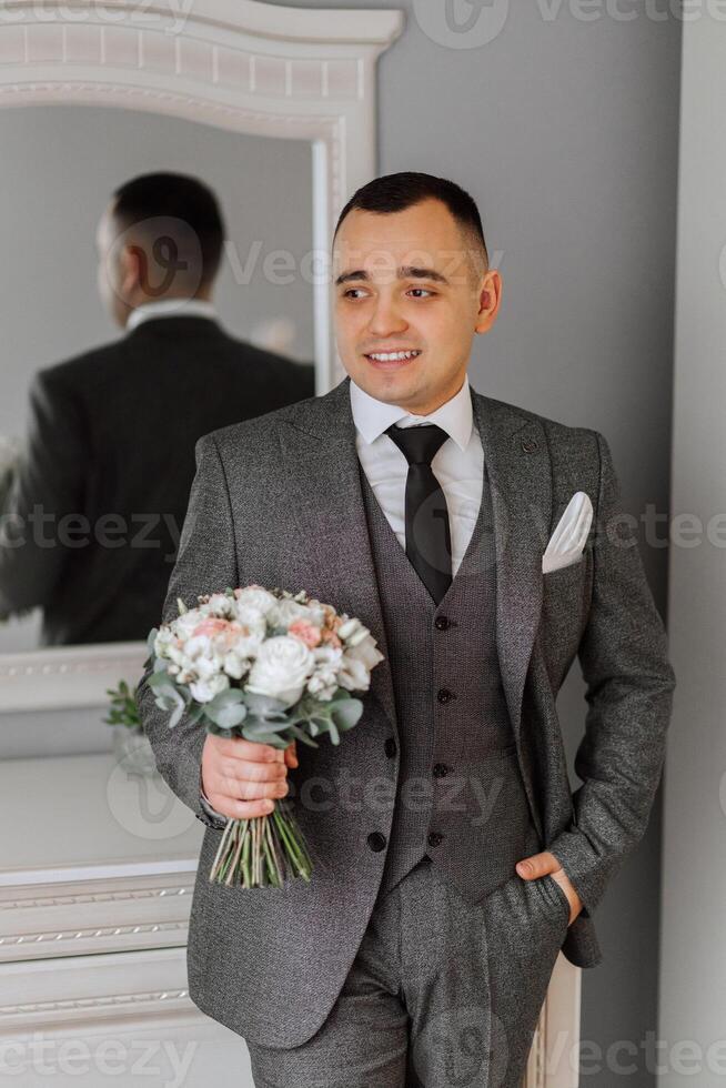 un' uomo nel un' grigio completo da uomo è Tenere un' mazzo, in posa nel il suo camera. ritratto di il sposo. moda e stile. attività commerciale foto