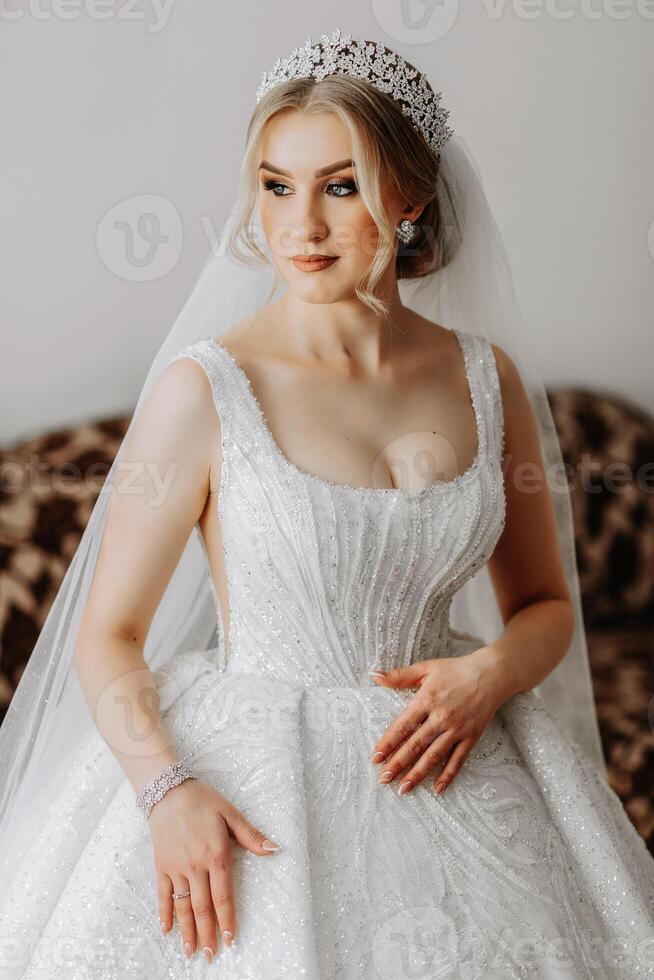 il sposa nel un' bianca vestire, velo pose nel bellissimo illuminazione nel sua camera. mattina di il sposa. un' bellissimo tiara foto