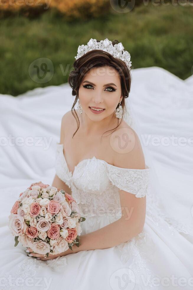 brunetta sposa nel fuori dalla spalla pizzo bianca vestito e tiara in posa con un' mazzo di bianca e rosa fiori mentre seduta su verde erba. bellissimo capelli e trucco. primavera nozze foto