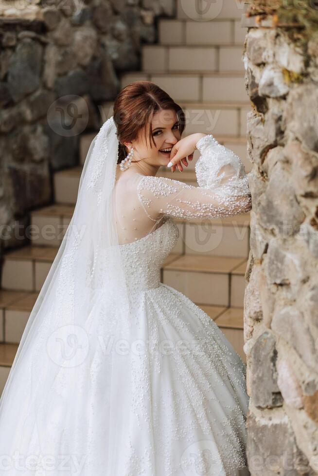 il sposa nel un' magnifico vestito e velo, in posa vicino un' pietra parete. magnifico vestito con lungo maniche, Aperto rompersi. estate nozze foto