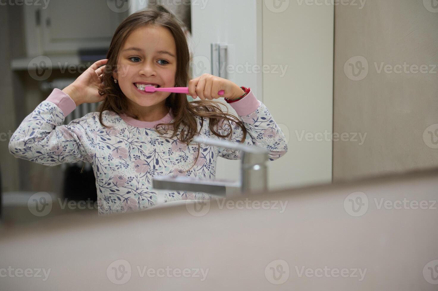 riflessione nel il bagno specchio di un' carino bambino ragazza spazzolatura denti, vestito nel pigiama. dentale igiene e orale cura foto