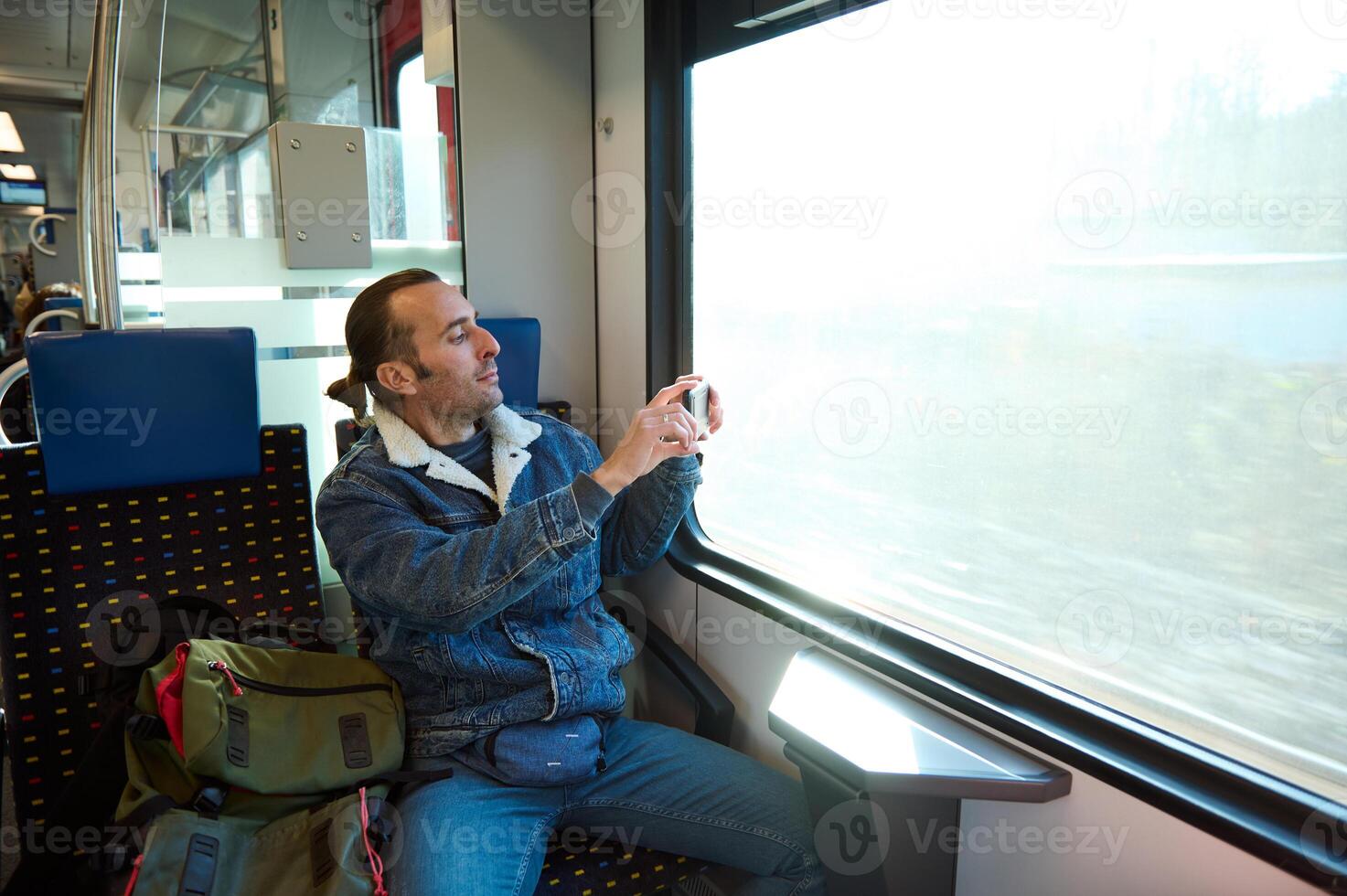 allegro giovane adulto caucasico uomo nel casuale denim, guardando su il finestra durante il suo mattina commutare su un urbano leggero rotaia treno, sorridente e assunzione foto di natura su il finestra su il suo smartphone