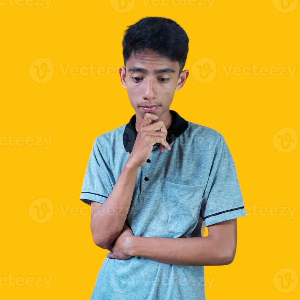 giovane riflessivo asiatico uomo Tenere mano su mento guardare pensieroso, indossare grigio maglietta su giallo sfondo. foto