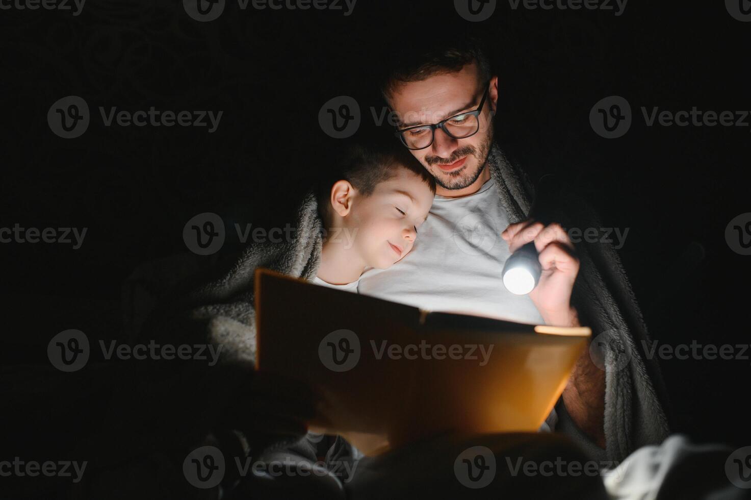 padre e figlio con torcia elettrica lettura libro sotto coperta a casa. foto