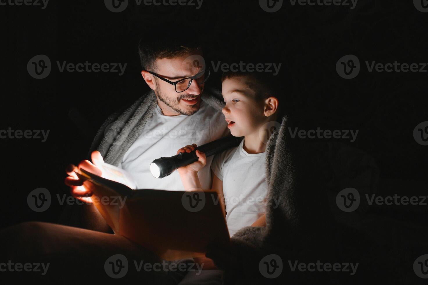 padre e figlio con torcia elettrica lettura libro sotto coperta a casa. foto