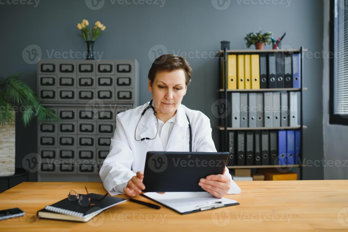 ritratto tiro di mezzo anziano femmina medico seduta a scrivania e Lavorando nel ufficio. foto