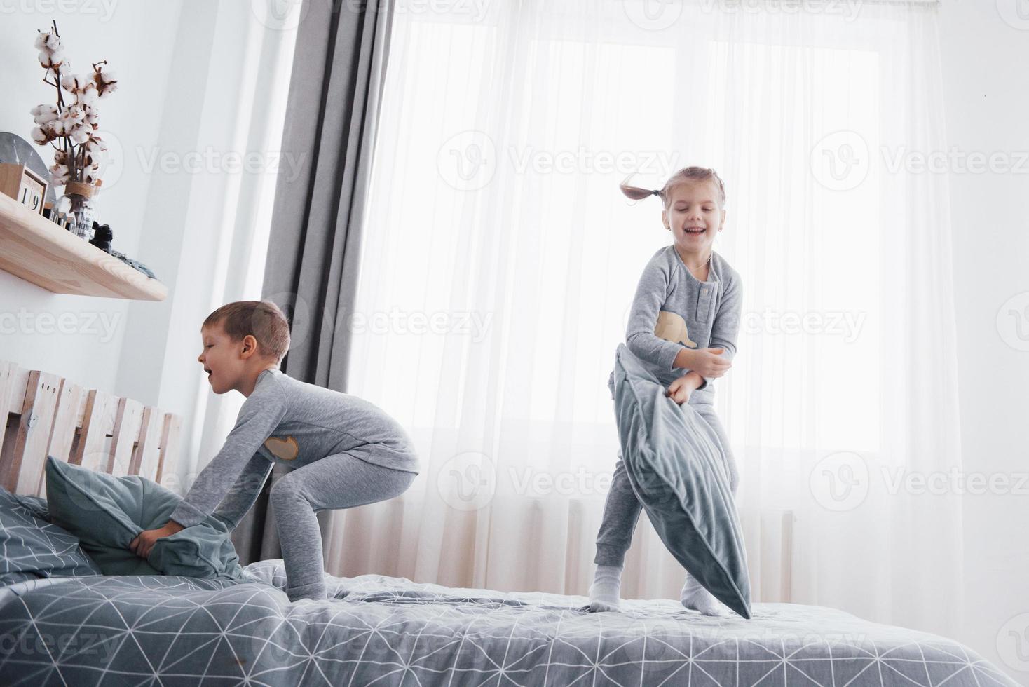 bambini felici che giocano nella camera da letto bianca. ragazzino e ragazza, fratello e sorella giocano sul letto indossando il pigiama. biancheria da notte e biancheria da letto per neonati e bambini. famiglia a casa foto