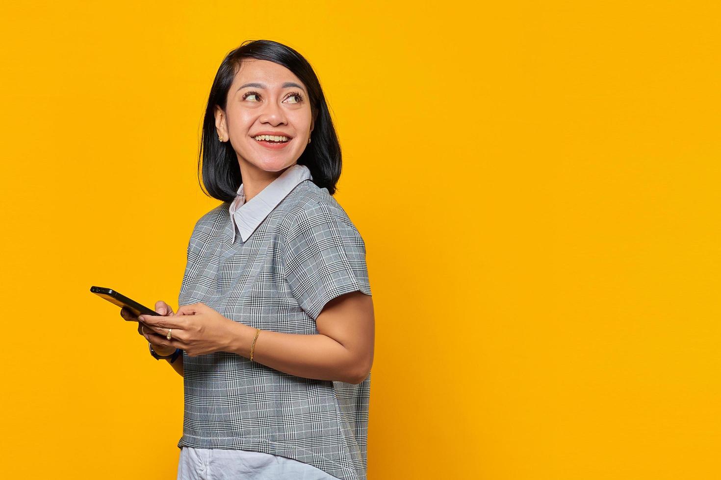 ritratto di giovane donna asiatica sorridente che tiene il telefono cellulare e guarda da parte su sfondo giallo foto