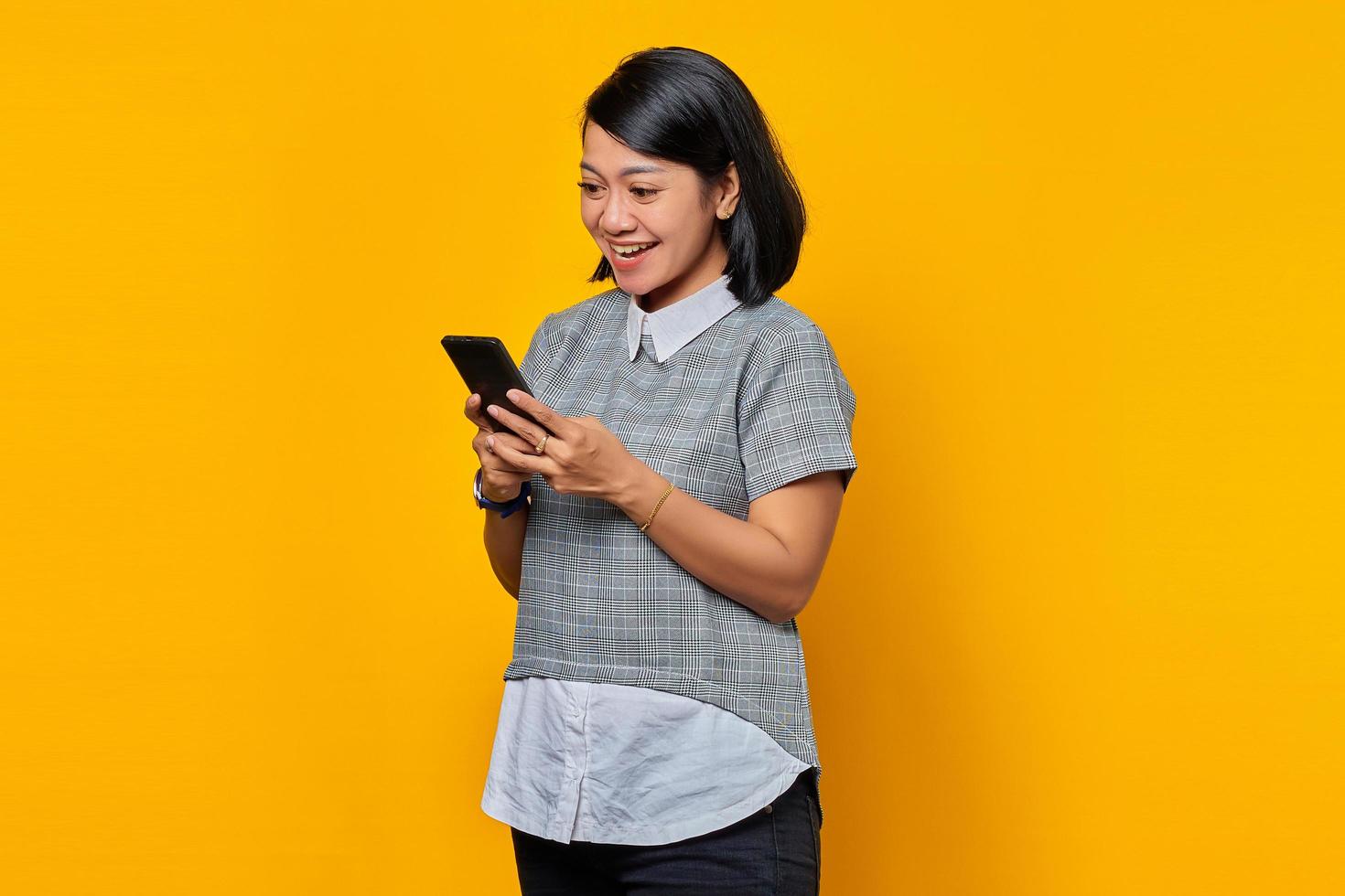 ritratto di allegra giovane donna asiatica guardando il messaggio sullo smartphone isolato su sfondo giallo foto