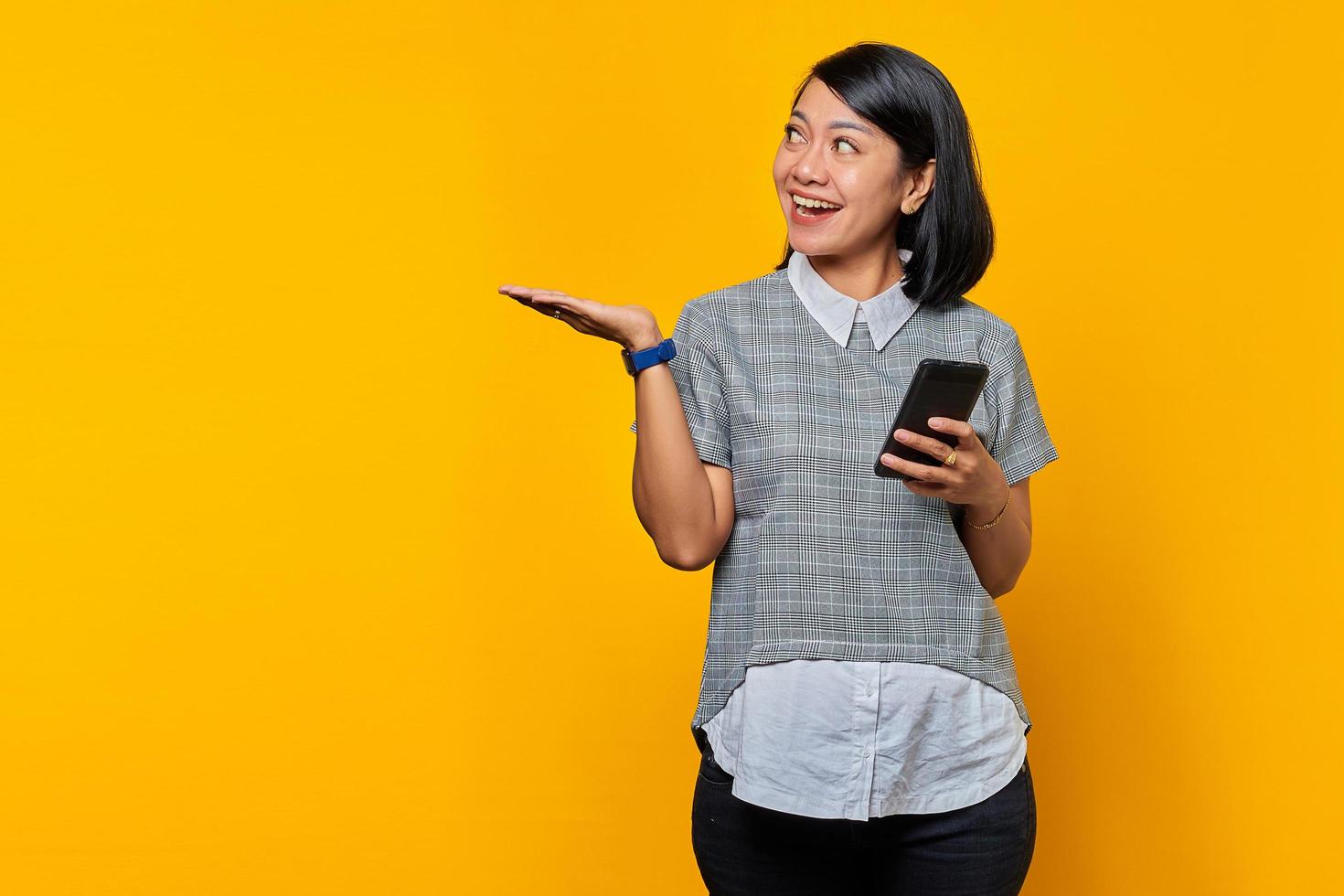 ritratto di una donna asiatica sorridente che guarda da parte con i palmi che mostra lo spazio vuoto e tiene il telefono cellulare su sfondo giallo foto