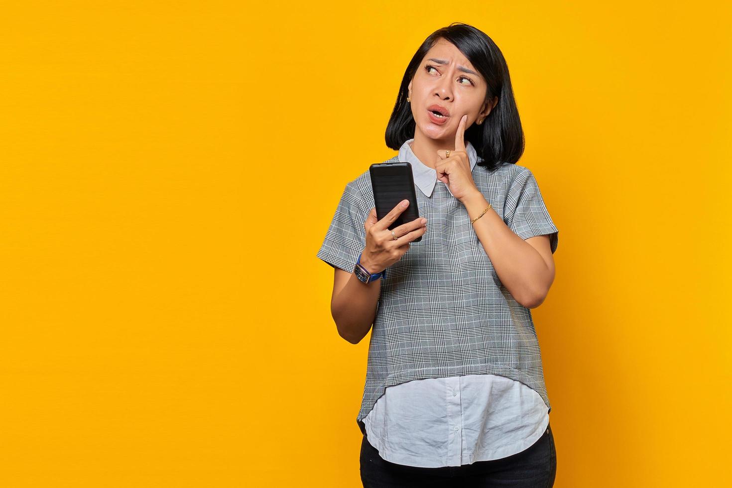 ritratto di una giovane donna asiatica sorpresa che guarda da parte pensando a una domanda con il dito sul mento e tiene il cellulare foto