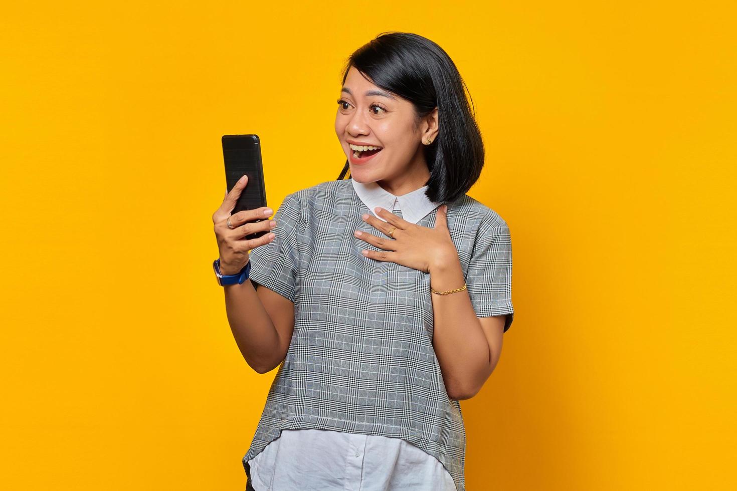 allegra giovane donna asiatica che guarda il telefono cellulare e tiene i palmi sul petto su sfondo giallo foto