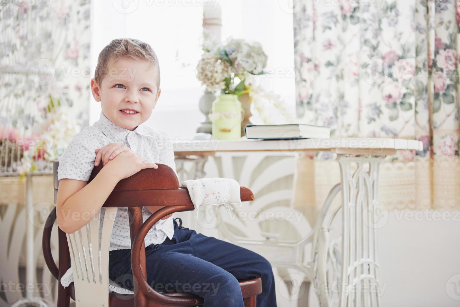 il ragazzino in pantaloni blu jeans è seduto sulla sedia di legno nella stanza foto