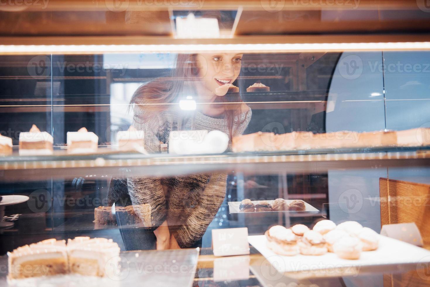 donna sorridente in telecamera attraverso la vetrina con dolci e torte in un moderno bar interno foto