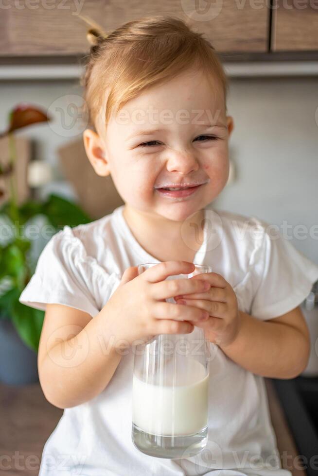 contento bambino ragazza seduta a il tavolo nel il cucina e potabile latte foto