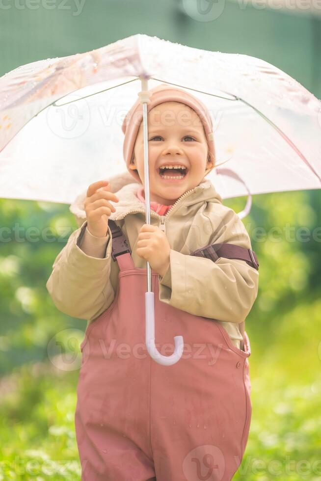 contento ridendo bambino ragazza 2-3 anno vecchio indossare impermeabile Abiti e Tenere rosa ombrello avere un' divertimento su casa Giardino dietro la casa nel piovoso giorno foto