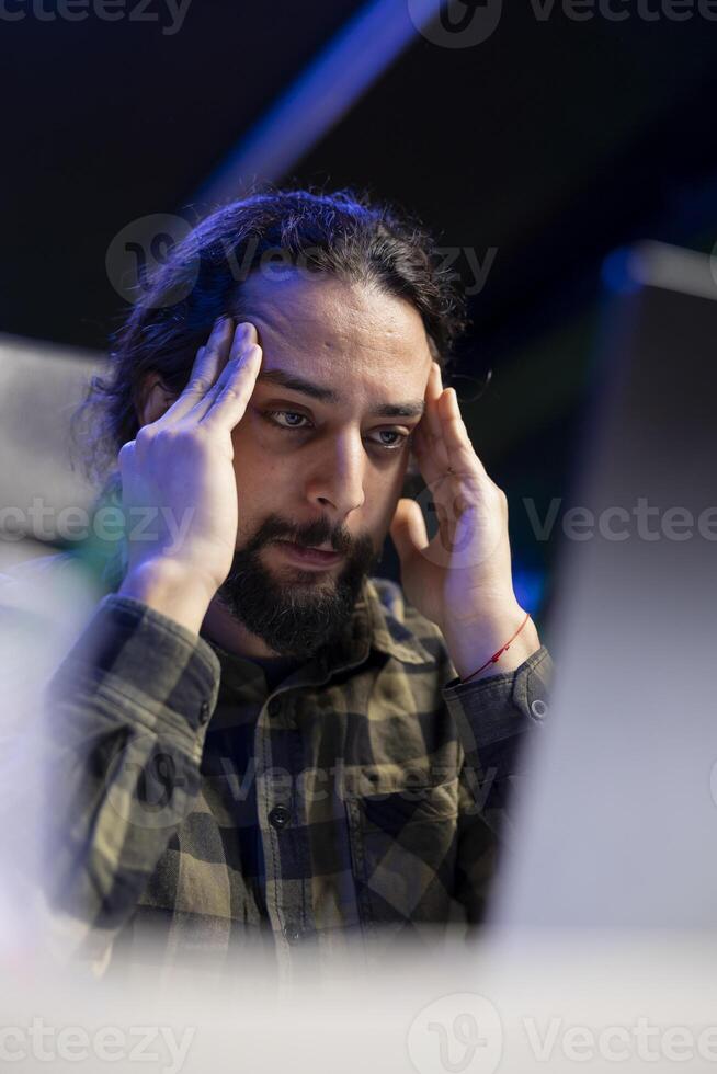 sensazione malato e esausto è un' maschio Università alunno seduta nel davanti di il suo il computer portatile. stressato persona alleviare mal di testa di sfregamento il suo templi mentre controllo il suo portatile computer. foto