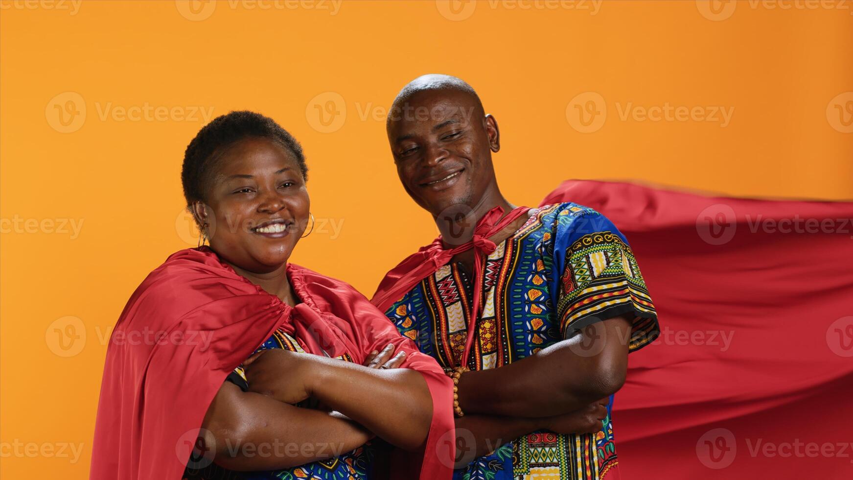 africano americano coppia in posa con supereroe rosso mantello, recitazione come potente grave eroi con comico libro mantelli. etnico persone in piedi nel studio con braccia attraversato, orgoglioso forte Modelli. foto