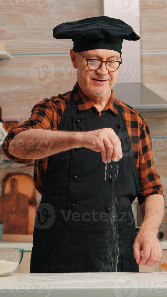 anziano panettiere nel casa cucina mettendo Grano Farina al di sopra di di legno tavolo. pensionato anziano capocuoco con bonete e grembiule aspersione, setacciare vagliatura crudo ingredienti di mano cottura al forno fatti in casa Pizza, pane. foto