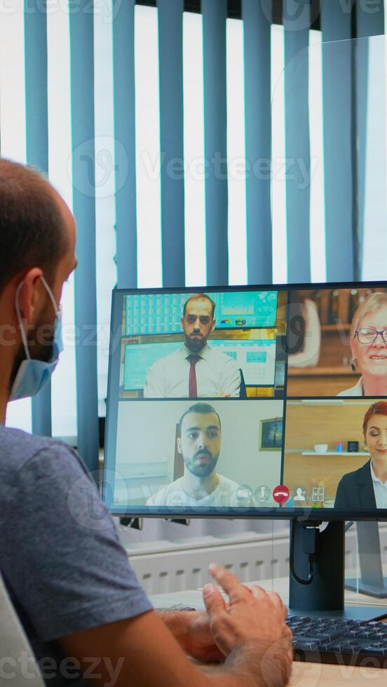 uomo con protezione maschera partecipando a in linea gruppo video conferenza nel nuovo normale ufficio. libero professionista Lavorando nel posto di lavoro chat parlando avendo virtuale incontro, utilizzando Internet tecnologia foto